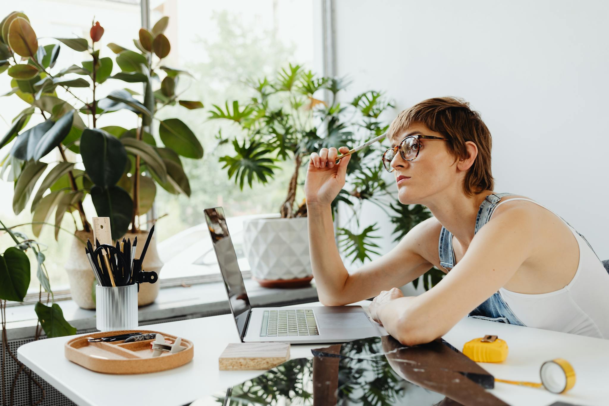 Woman in casual wear working on laptop at desk surrounded by plants. Creative and thoughtful workspace.